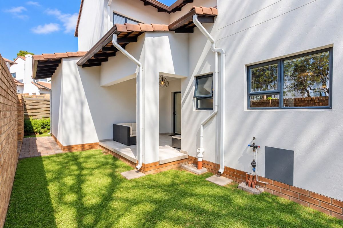 A well-maintained rear exterior view of the townhouse featuring a paved patio area, small lawn, and clean white facade with terracotta roof tiles.