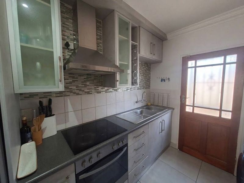 Compact kitchen featuring built-in cabinetry, a modern electric hob with extractor fan, stainless steel sink, and tiled backsplash with direct exterior access.