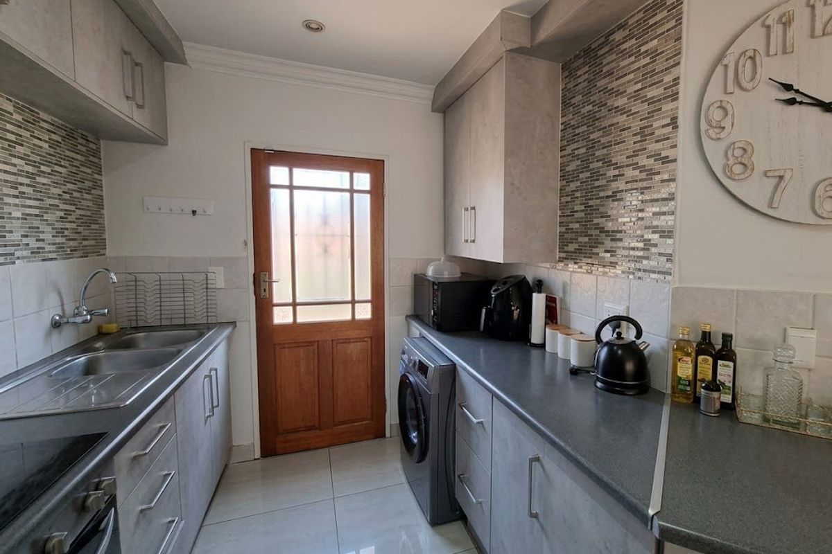 A functional galley-style kitchen featuring modern grey cabinetry, mosaic tile backsplashes, tiled flooring, and integrated laundry facilities with direct exterior access.
