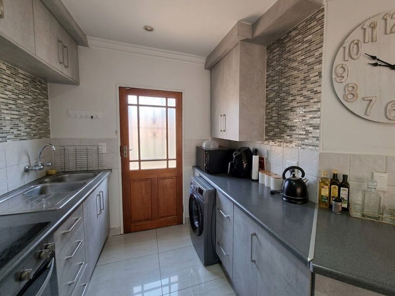 A functional galley-style kitchen featuring modern grey cabinetry, mosaic tile backsplashes, tiled flooring, and integrated laundry facilities with direct exterior access.