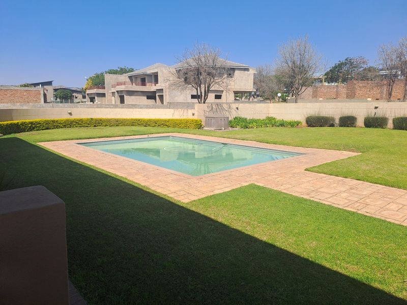 A well-maintained residential backyard featuring a rectangular swimming pool, paved surround, and established lawn enclosed by a perimeter boundary wall.