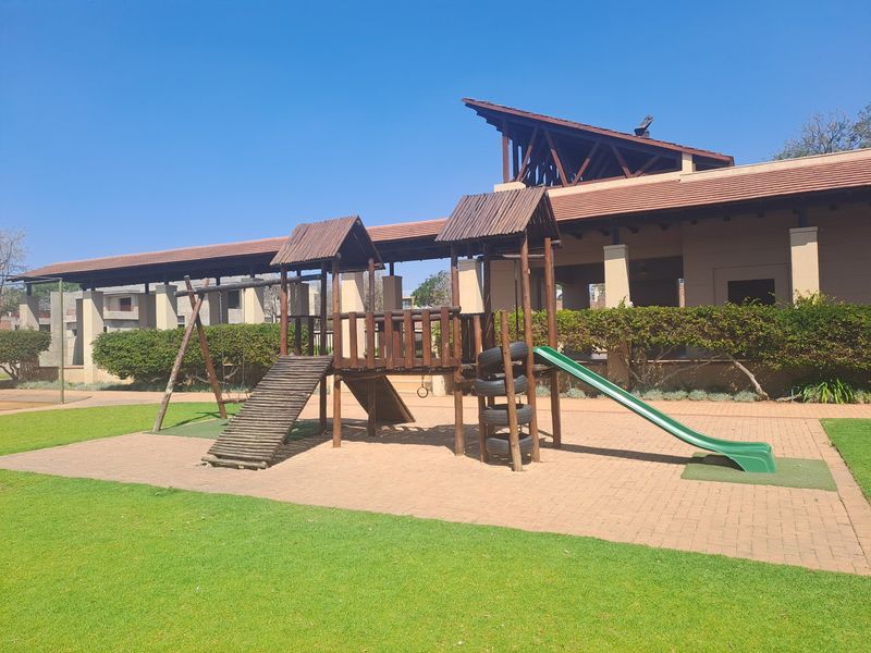 A well-maintained communal garden area featuring a wooden playground structure set on a paved surface with manicured lawn and established hedging.