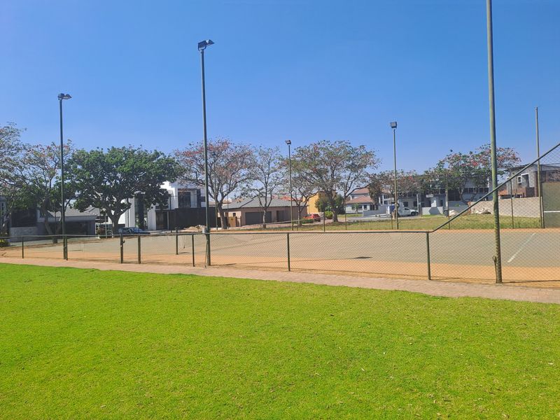 A well-maintained outdoor tennis court featuring chain link fencing, floodlight poles, and an adjacent manicured lawn area within a residential estate.