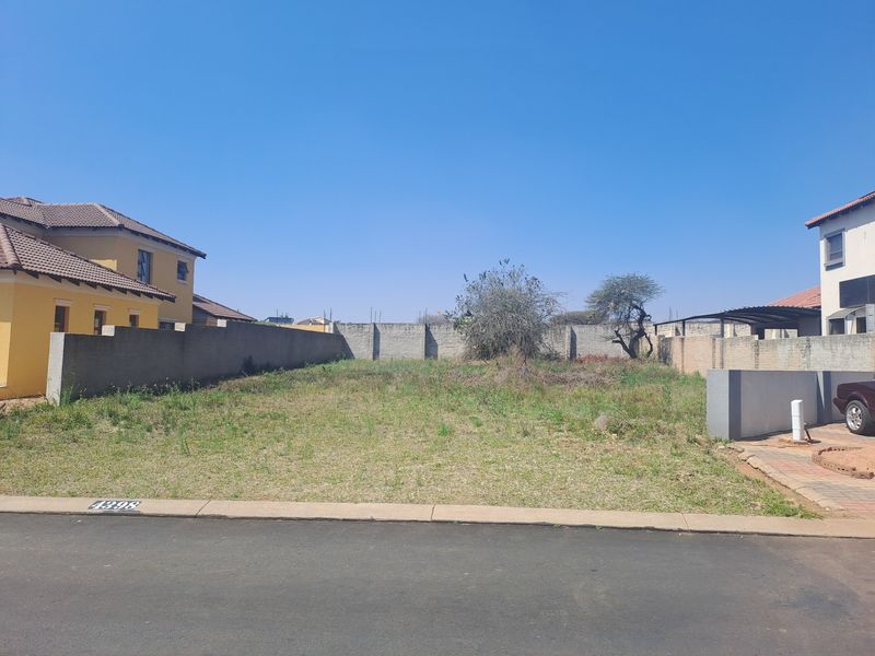 A vacant residential plot situated between existing homes, featuring a perimeter boundary wall and street-level access.