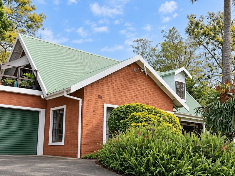 A charming brick house featuring a green metal roof, a roller garage door, and an elevated balcony set within a lush, landscaped garden.