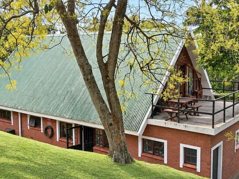 A rear view of a brick-built A-frame house featuring a green corrugated metal roof and an elevated balcony with metal railings.