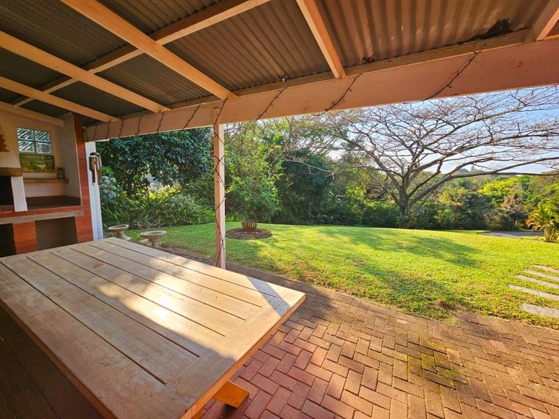 Covered outdoor patio featuring a built-in brick braai, timber roof structure, and brick paving overlooking a spacious garden.