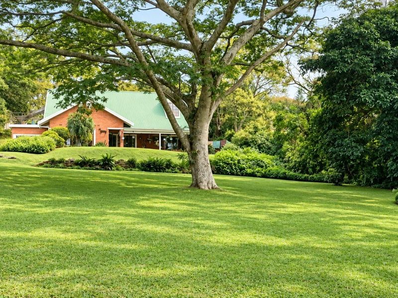 A spacious, well-maintained garden featuring a large lawn, mature trees, and a view of the brick house with a metal roof.