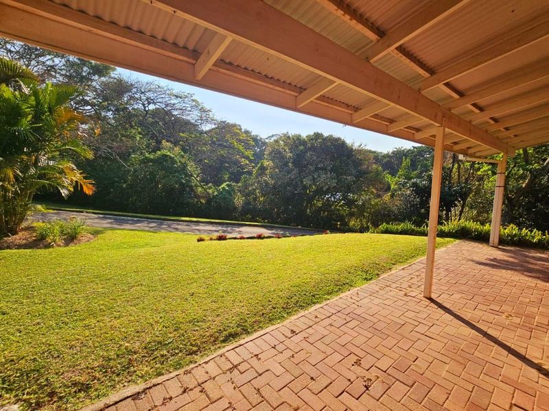 A covered outdoor patio featuring brick paver flooring and timber support posts, overlooking a lush green garden and driveway.