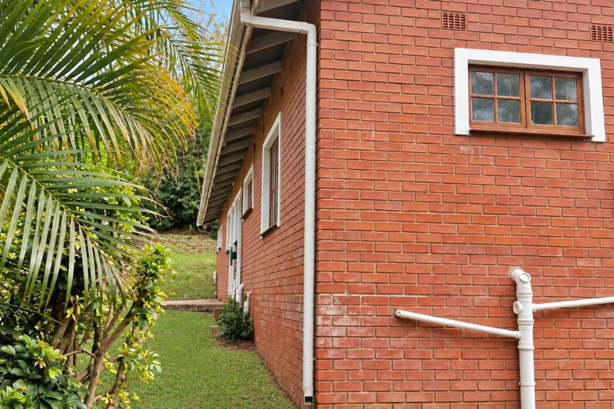 Side view of the property showing exposed red brick exterior, timber-framed windows, and external plumbing infrastructure set against a garden area.