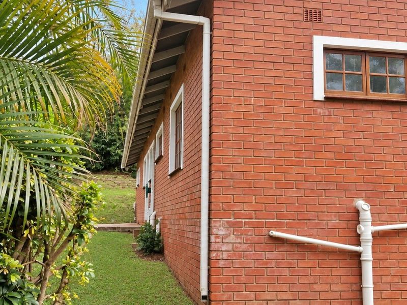 Side view of the property showing exposed red brick exterior, timber-framed windows, and external plumbing infrastructure set against a garden area.