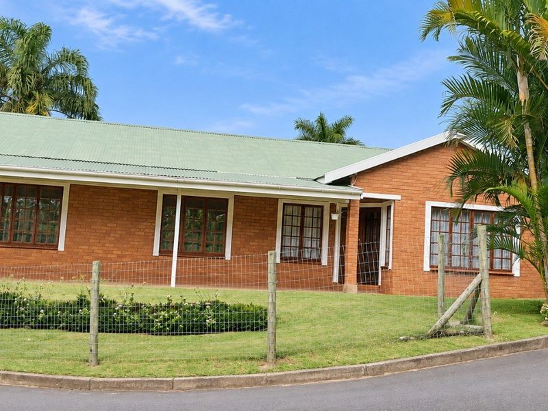 A single-story brick house with a green corrugated metal roof, timber-framed windows, and a front lawn enclosed by wire mesh fencing.