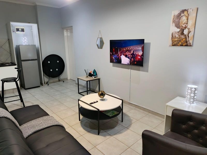 An open-plan living area featuring tiled flooring, a compact kitchen corner, and a wall-mounted television, creating a functional and modern living space.