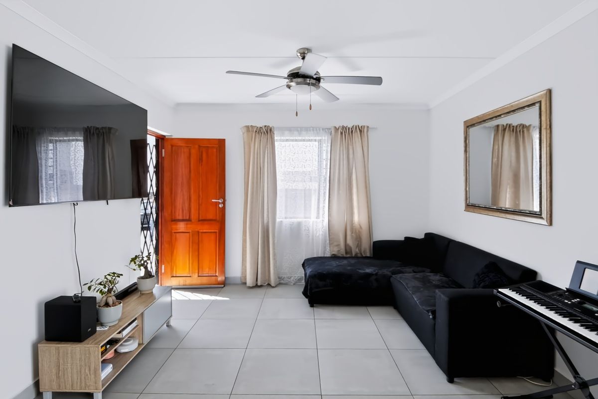 A bright and well-maintained living room featuring neutral tiled flooring, a ceiling fan, and direct access to the exterior via a timber entrance door.