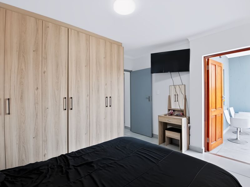 A well-maintained bedroom featuring extensive built-in light wood-grain cupboards, tiled flooring, and direct access to an adjoining room through a timber door.