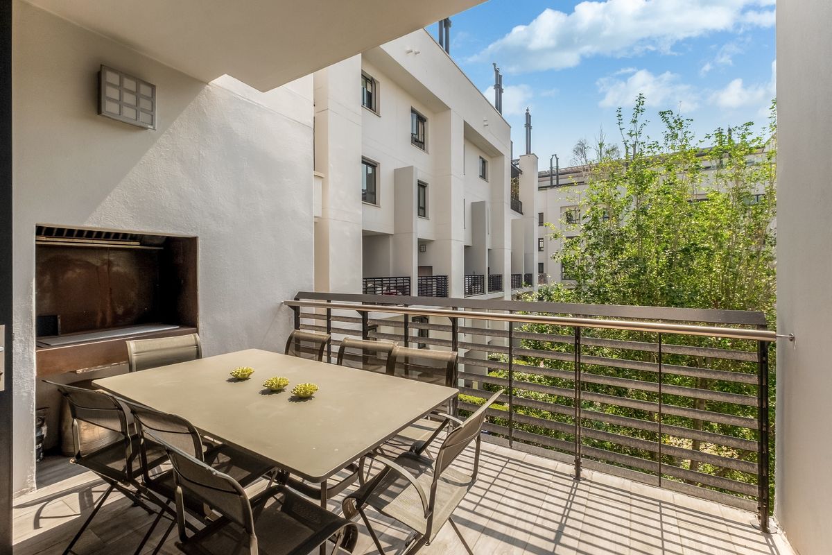 A spacious covered balcony featuring a built-in braai, metal balustrade, and tiled flooring, overlooking the apartment complex courtyard.