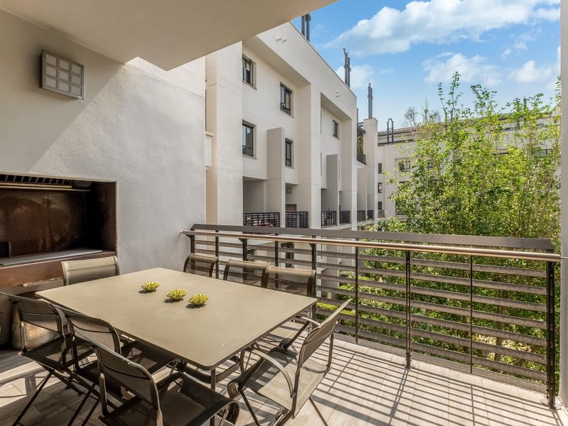 A spacious covered balcony featuring a built-in braai, metal balustrade, and tiled flooring, overlooking the apartment complex courtyard.