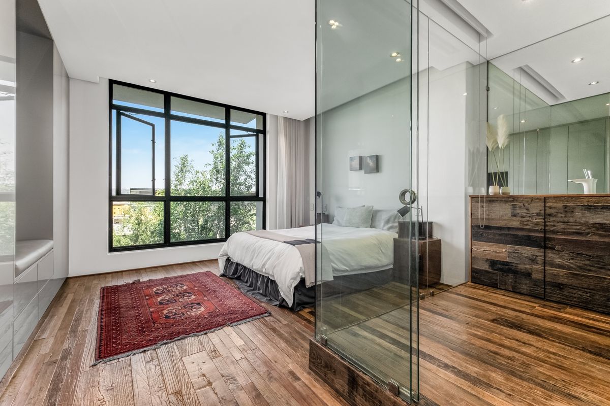Modern bedroom featuring rich hardwood flooring, large black-framed windows, and a contemporary glass-enclosed ensuite bathroom with integrated cabinetry.