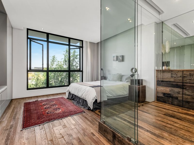 Modern bedroom featuring rich hardwood flooring, large black-framed windows, and a contemporary glass-enclosed ensuite bathroom with integrated cabinetry.