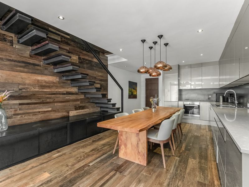 Modern open-plan kitchen and dining area featuring a striking floating staircase, reclaimed wood feature wall, high-gloss cabinetry, and premium stone countertops.