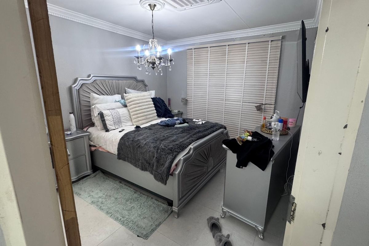 A well-maintained bedroom featuring tiled flooring, decorative coving, a central chandelier with a ceiling rose, and fitted venetian blinds.