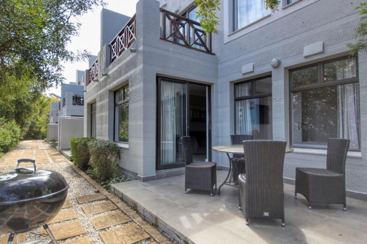 A ground-floor patio area featuring tiled flooring, sliding door access to the interior, and a timber-railed balcony above, set against a landscaped exterior.