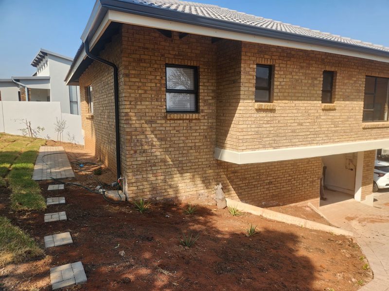 Rear view of a face brick house featuring a tiled roof, aluminium window frames, and a paved walkway leading through an unfinished garden area.