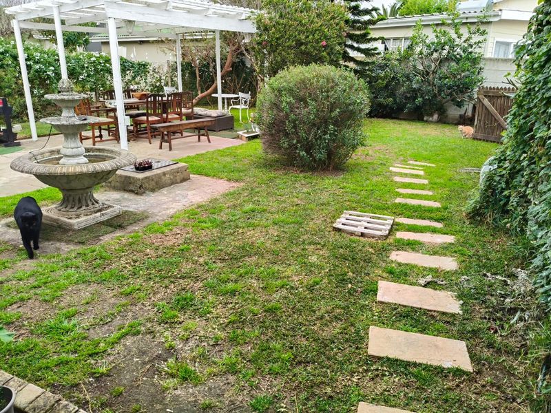 A rear garden featuring a white pergola, a stone fountain, a paved patio area, and a stepping stone path through the lawn.