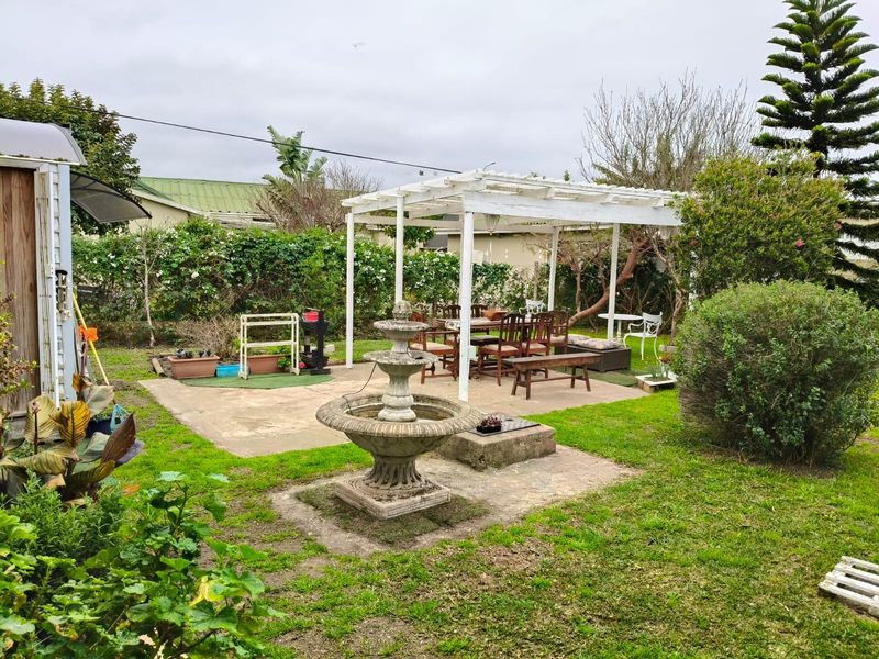 A rear garden featuring a concrete patio area with a white pergola structure and a central stone fountain, surrounded by established landscaping.