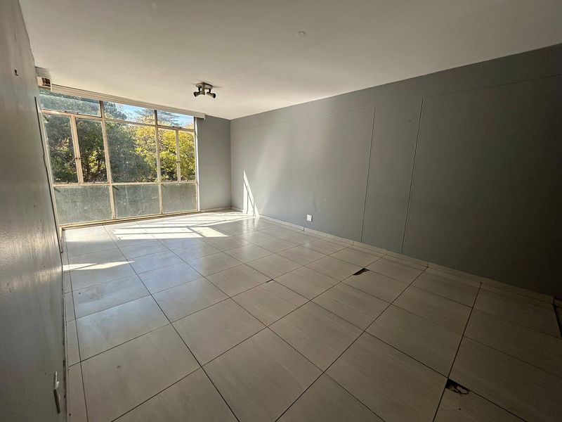 An empty living room featuring large windows and tiled flooring, requiring some maintenance to the floor tiles.