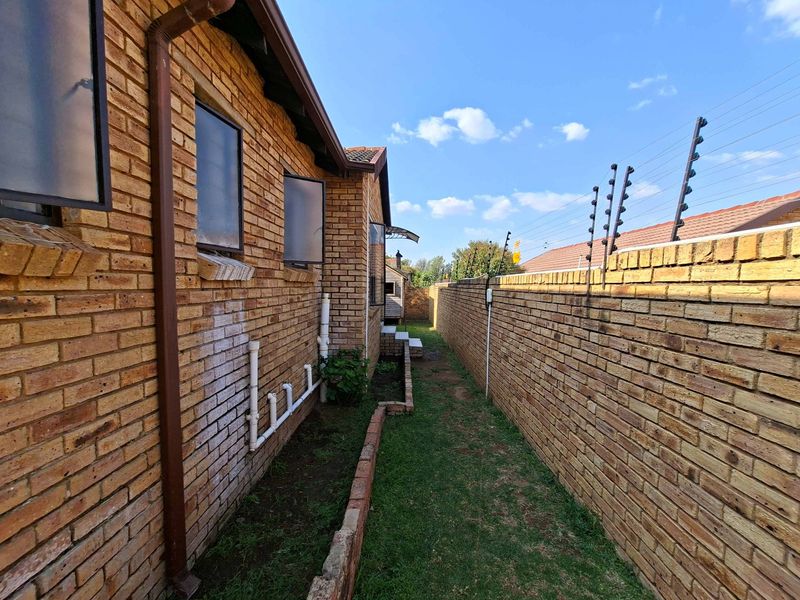 A side exterior view of the townhouse featuring face brick walls, a narrow garden strip, and perimeter boundary walls with electric fencing.