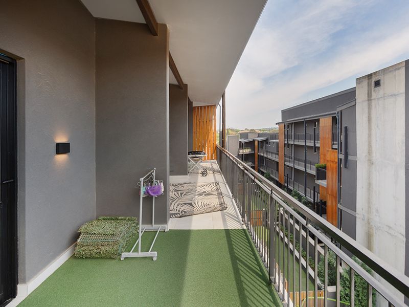 A spacious covered balcony featuring a metal balustrade, artificial grass flooring, and wall-mounted lighting, offering an open view of the apartment complex.