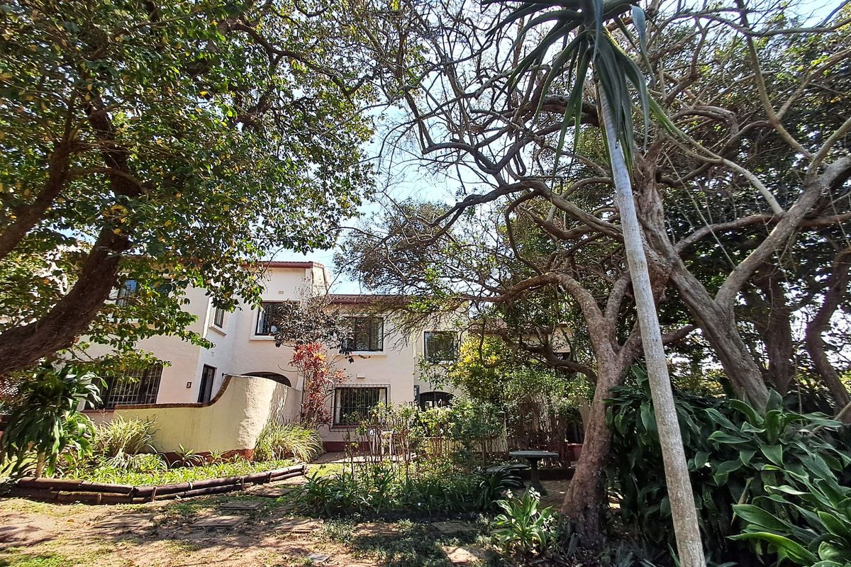 Rear view of the property featuring a landscaped garden with mature trees and a paved patio area adjacent to the building facade.