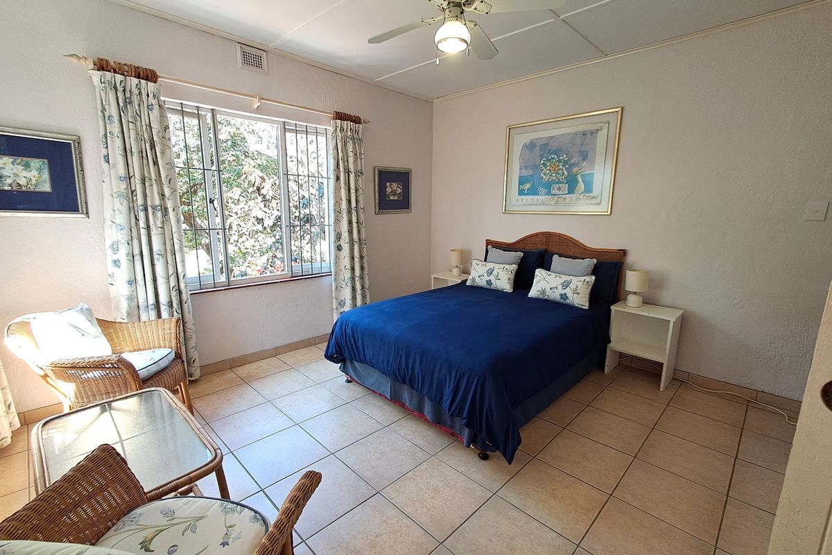 A spacious bedroom featuring neutral tiled flooring, a ceiling fan, and a large window with security bars, offering a functional and well-lit living space.