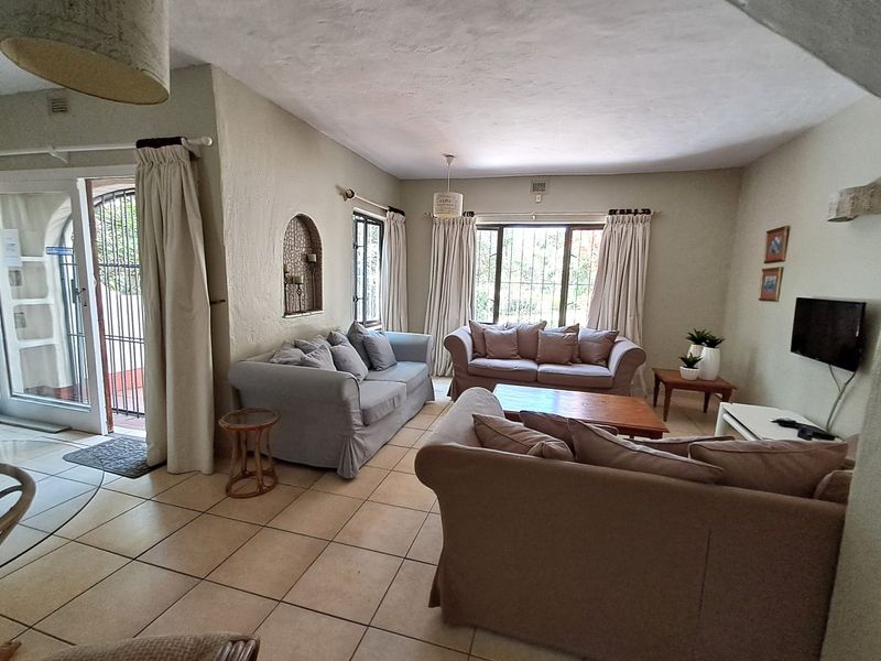 A comfortable living room featuring tiled flooring, an arched wall niche, and large windows with security bars, providing access to the exterior.