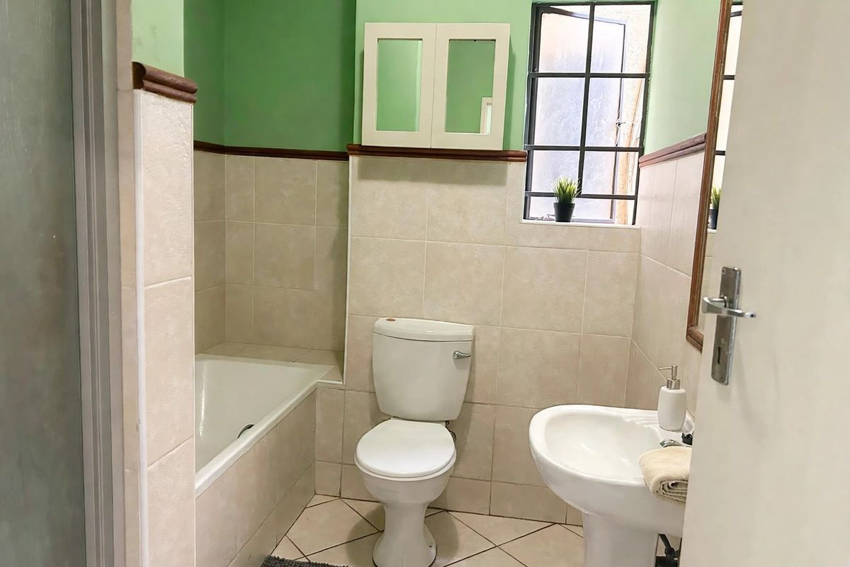 A functional bathroom featuring a bathtub, pedestal basin, and toilet, with neutral wall tiling and a window providing natural light.