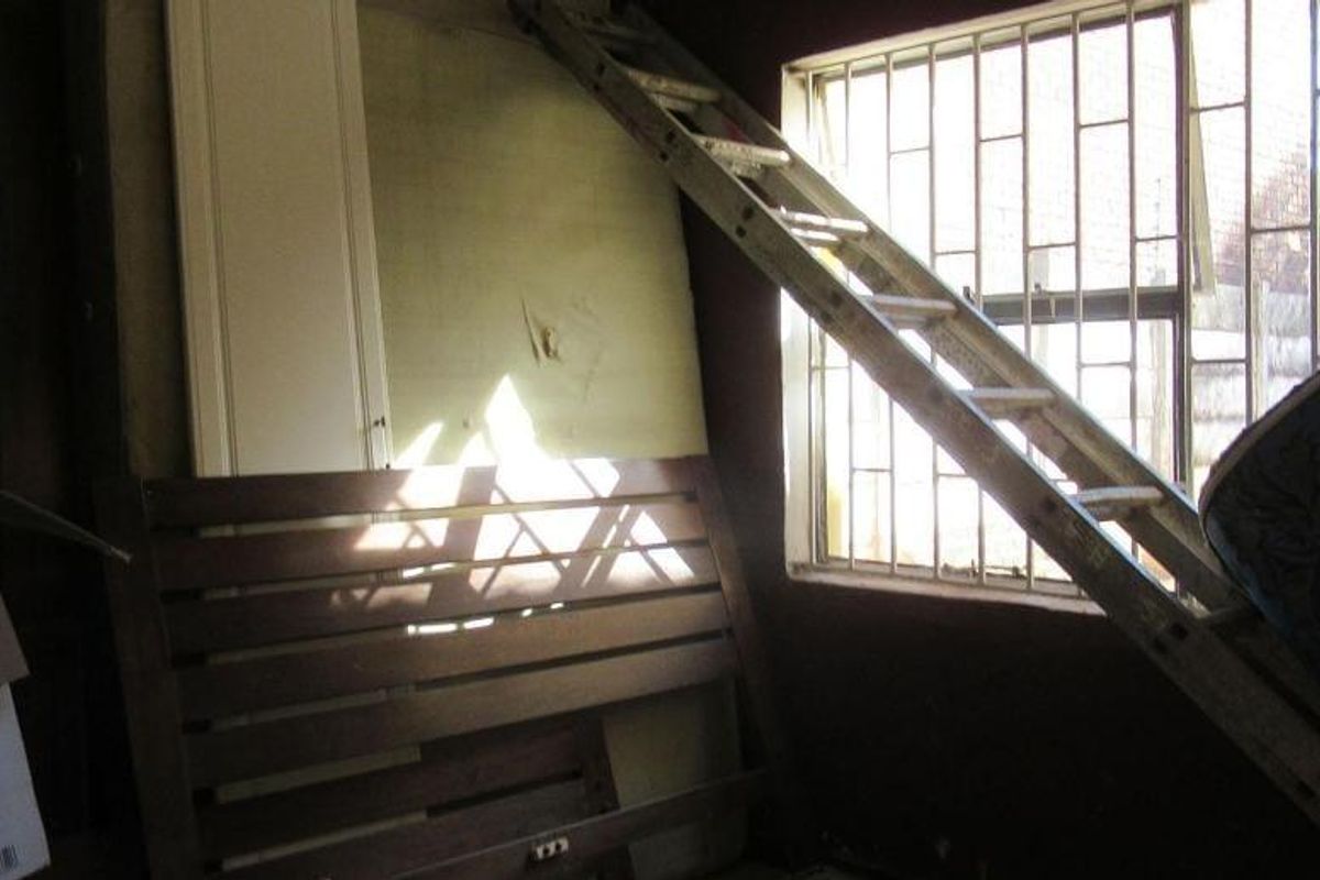 A bedroom space featuring a window with security bars and a built-in cupboard, currently in need of cosmetic refurbishment and wall repairs.