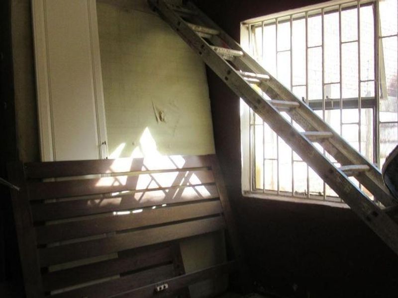 A bedroom space featuring a window with security bars and a built-in cupboard, currently in need of cosmetic refurbishment and wall repairs.