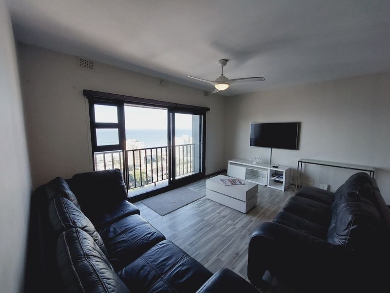 A comfortable living room featuring laminate flooring, a ceiling fan, and large sliding glass doors that open to a balcony with ocean views.