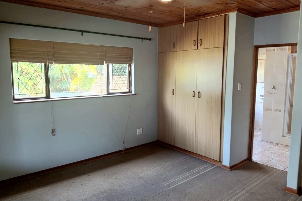 A vacant bedroom featuring built-in cupboards, a timber-clad ceiling, and a window with security bars, leading to an en-suite bathroom.