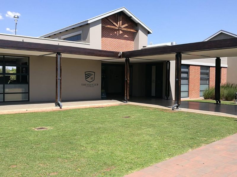 Front entrance view of The Els Club featuring a well-maintained lawn, covered walkway with timber beams, and modern brick and plaster facade.