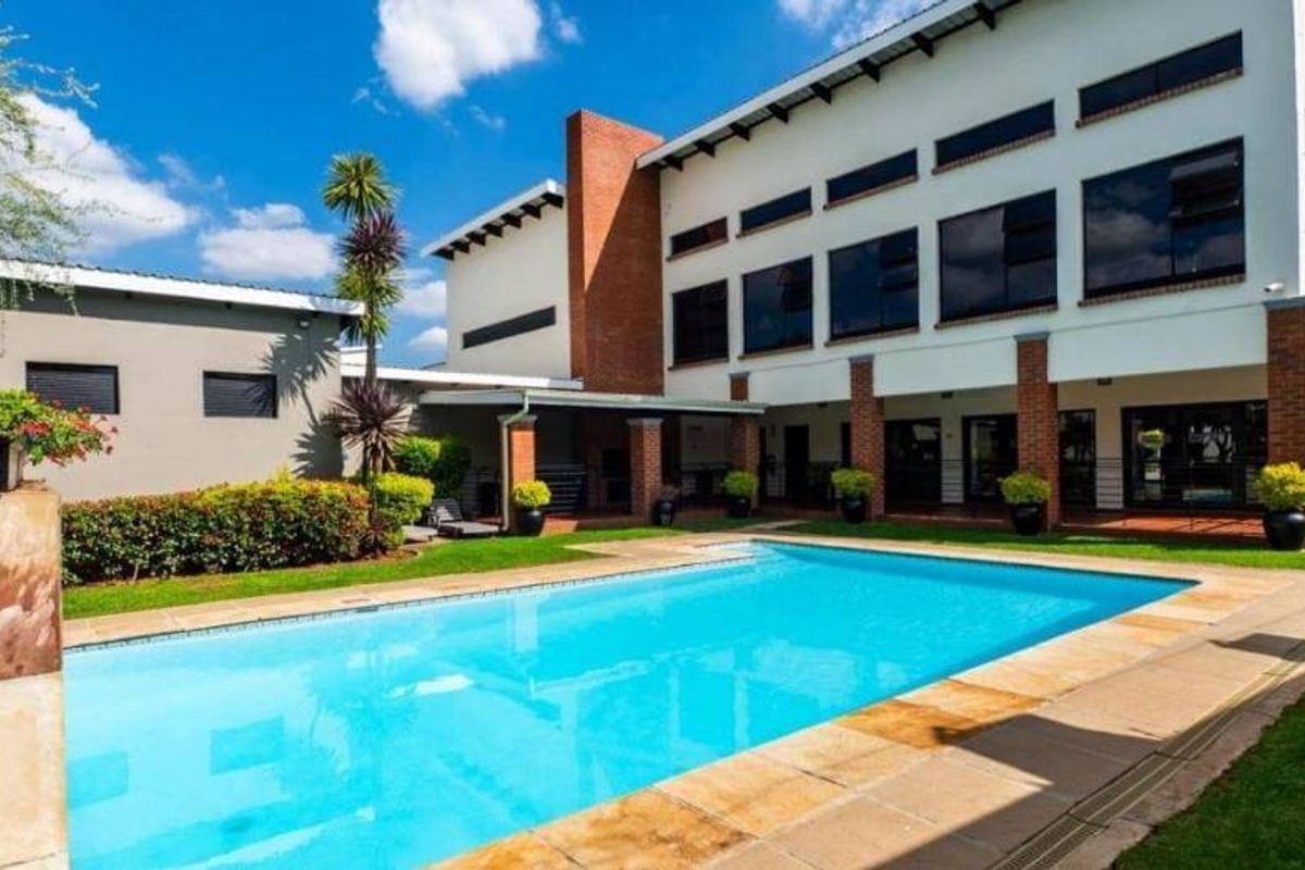 A well-maintained outdoor pool area featuring a clear swimming pool, paved surround, and a covered patio area set against a modern brick and plaster building.