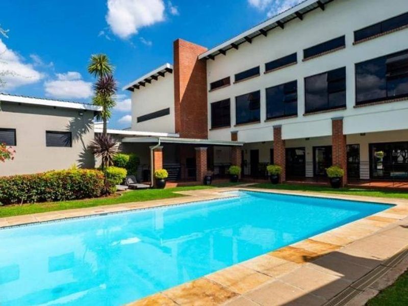 A well-maintained outdoor pool area featuring a clear swimming pool, paved surround, and a covered patio area set against a modern brick and plaster building.