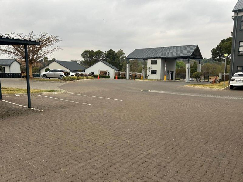 View of the apartment complex entrance featuring a security gatehouse, paved driveway, and covered parking bays.