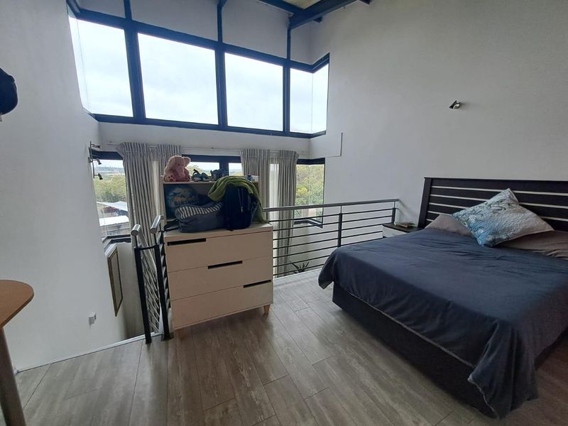 A spacious loft-style bedroom featuring double-volume windows, modern laminate flooring, and a metal balustrade overlooking the lower level.