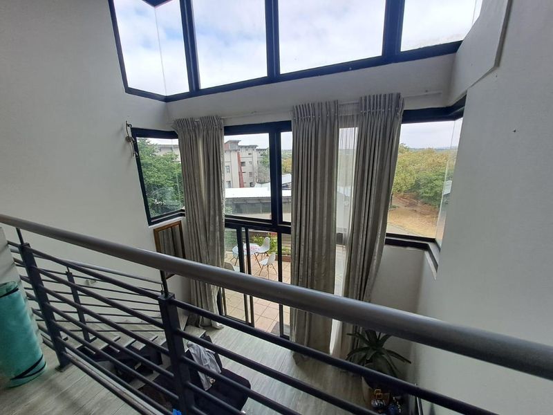 A bright, double-volume living area featuring expansive floor-to-ceiling windows, a modern metal balustrade, and direct access to an outdoor balcony.
