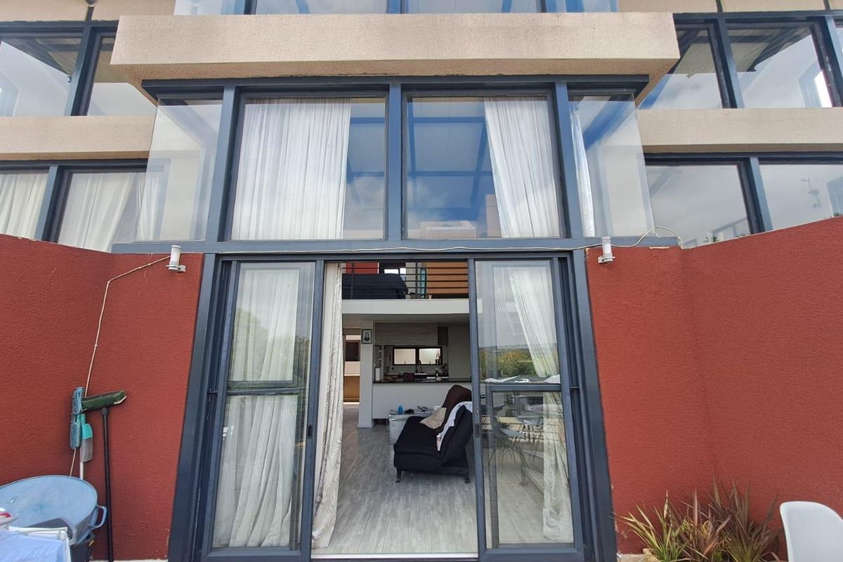 Rear exterior view of a modern apartment featuring a double-volume glass facade, large sliding doors, and a tiled patio area.