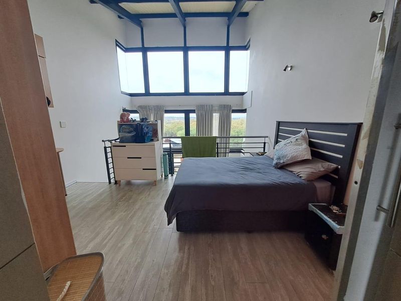 Spacious loft-style bedroom featuring high ceilings with exposed beams, large windows providing natural light, and modern laminate flooring.