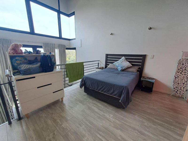 Spacious mezzanine bedroom featuring high ceilings, large windows with natural light, and modern laminate flooring overlooking the lower level.