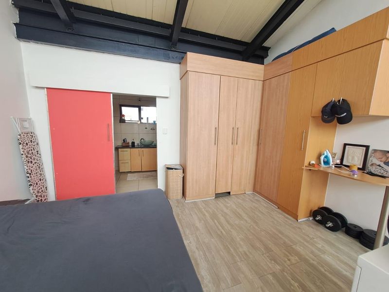 Bedroom featuring built-in wooden cupboards, exposed ceiling beams, and laminate flooring, with an adjacent view into a small kitchenette area.